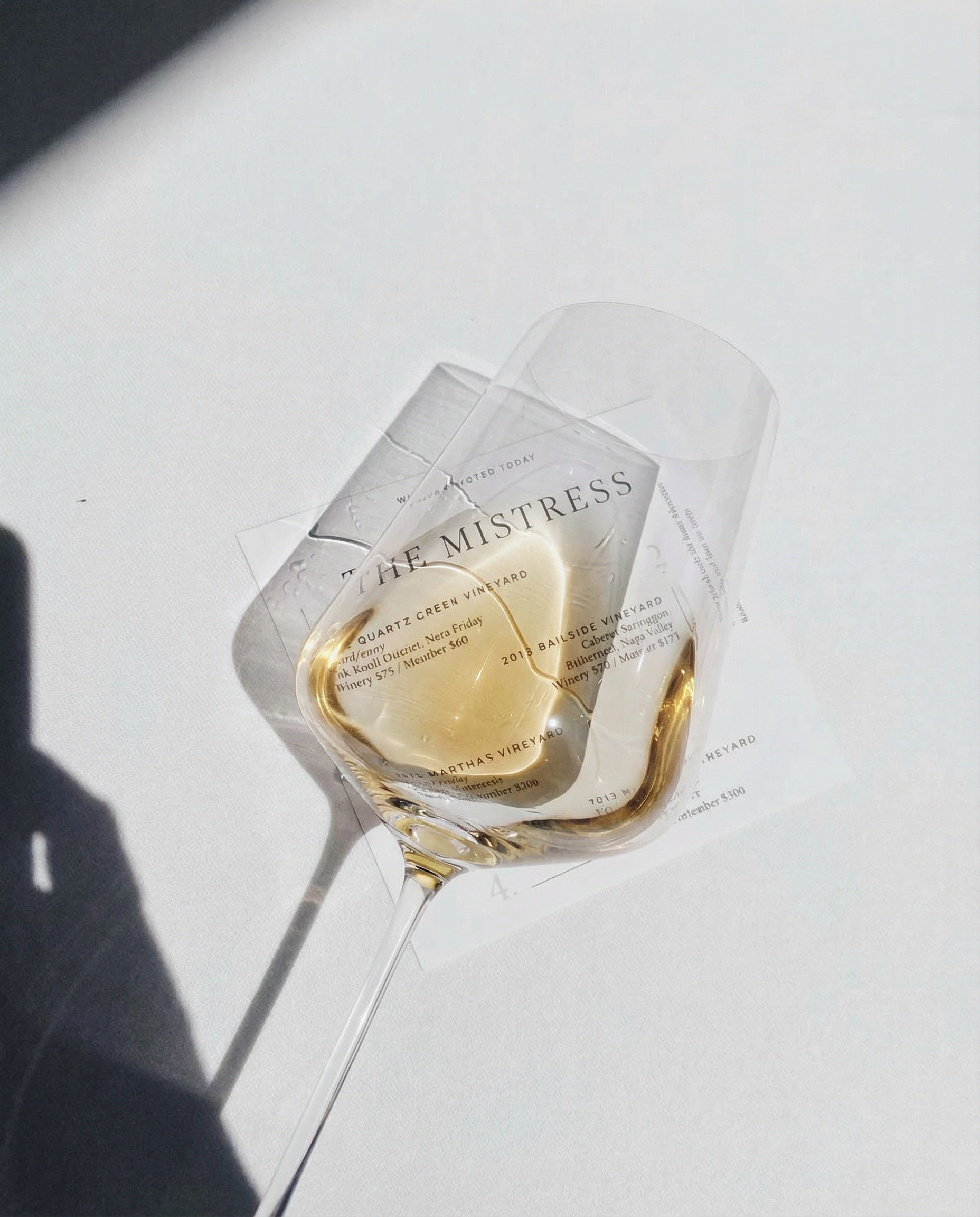 A clear wine glass labeled “The Mistress” lies on its side in bright sunlight, casting shadows while golden white wine glows inside the bowl over a printed tasting card.