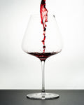 "The Mistress" red Wine Glass in white background and black - Luxury, premium and silent excellence design for wine lovers with poured red wine in it.  It has tapered big bowl for aromatic wine experience. Red Wine is pouring from above showing water fall effect and it causing bubbles in the wine!