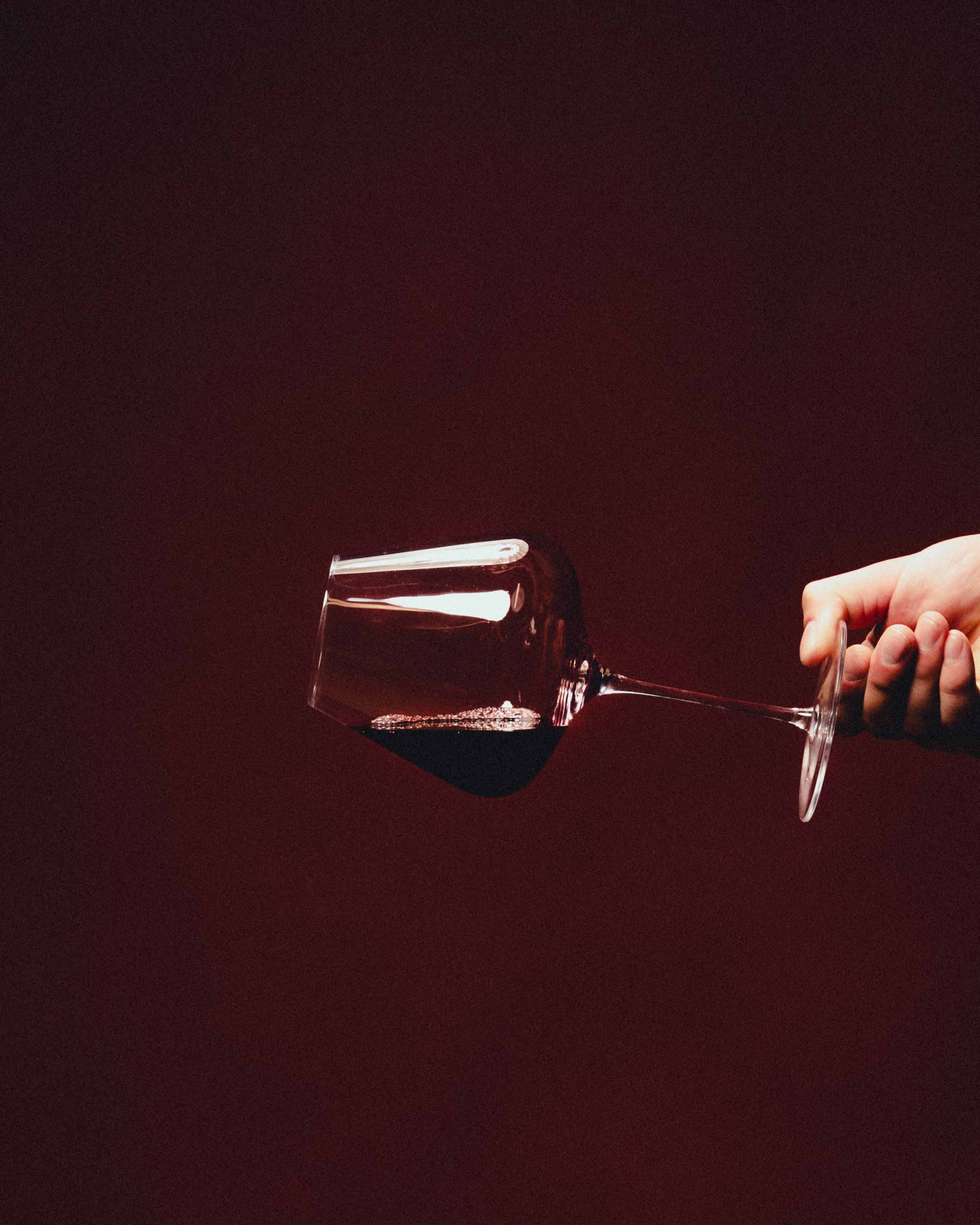 Kvsche Temptress red wine glass held sideways by hand against a dark burgundy studio backdrop